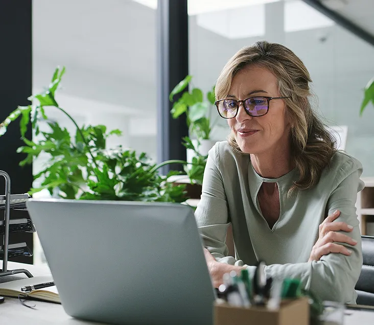 Confident woman wearing glasses and a light green blouse working at a laptop in a modern office with indoor plants.