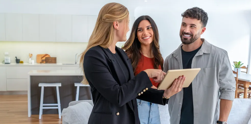 Female real estate agent showing a young couple something on a tablet in a modern kitchen-living space.
