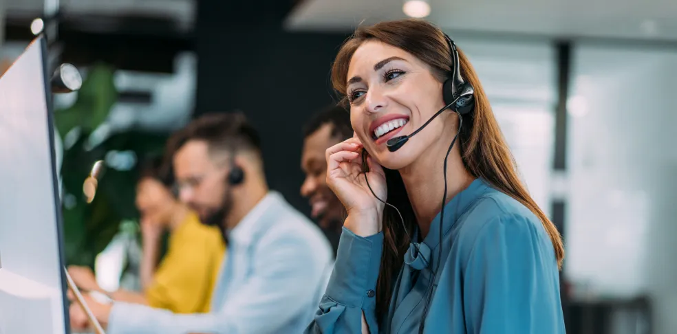 Smiling female customer service representative wearing a headset, with team members working in the background.