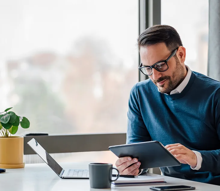 Man wearing glasses and a blue jumper using a tablet at a desk with a laptop, coffee mug, and potted plant in a bright office space.