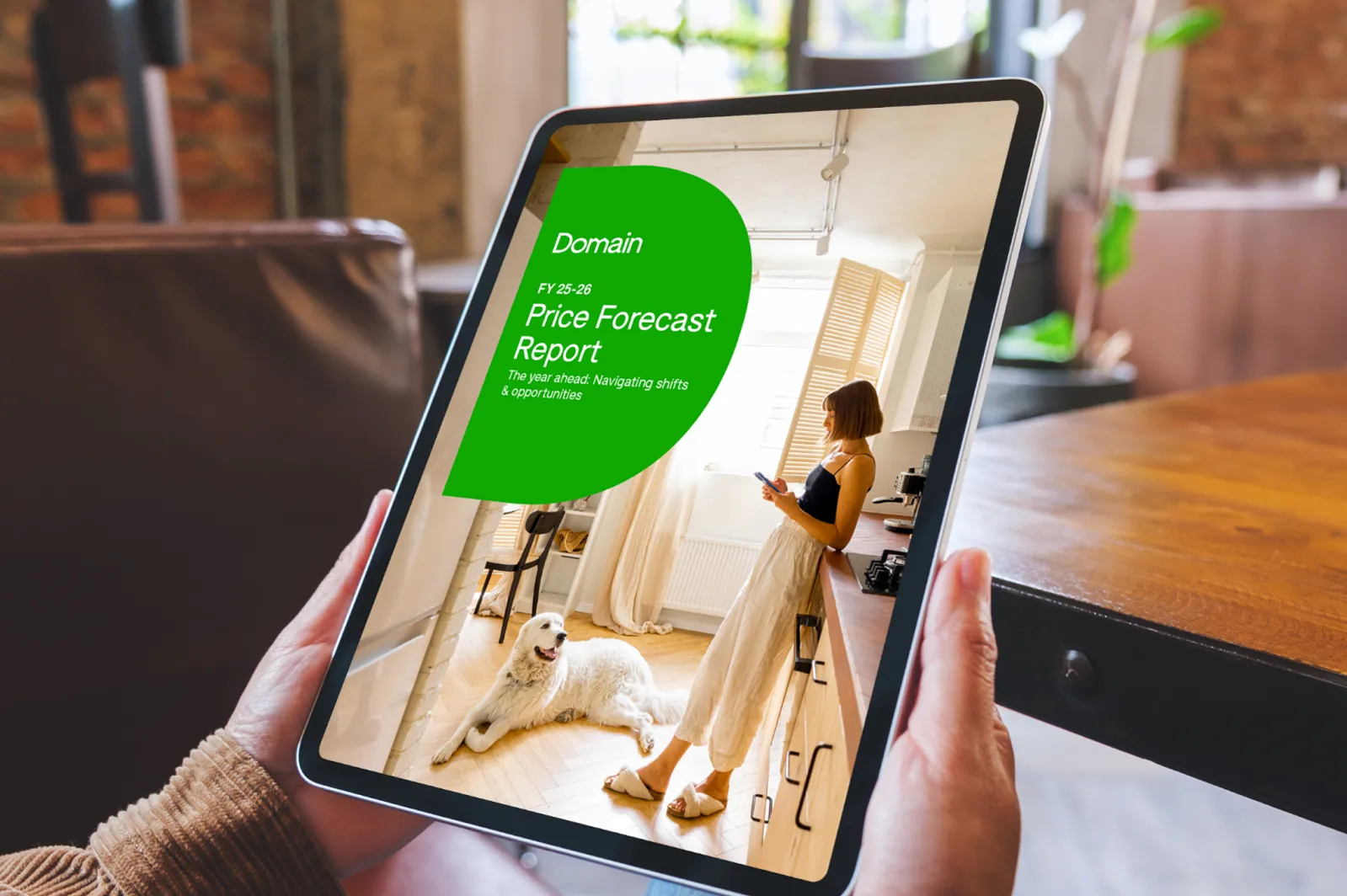 Person holding a tablet displaying the Domain FY25–26 Price Forecast Report, with a cover image of a woman standing in a sunlit kitchen next to a large white dog.