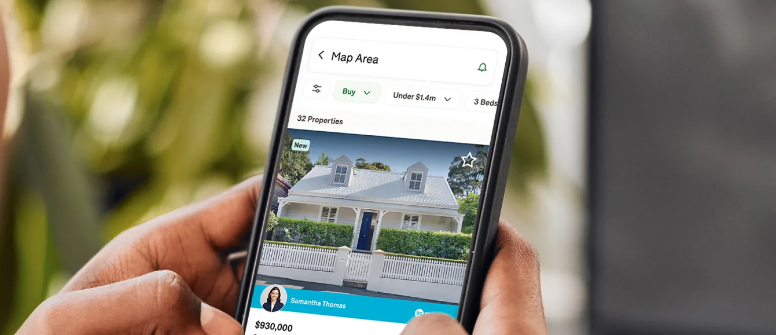 Person holding a smartphone displaying a real estate app with a property listing, showing a house for sale with filters applied.