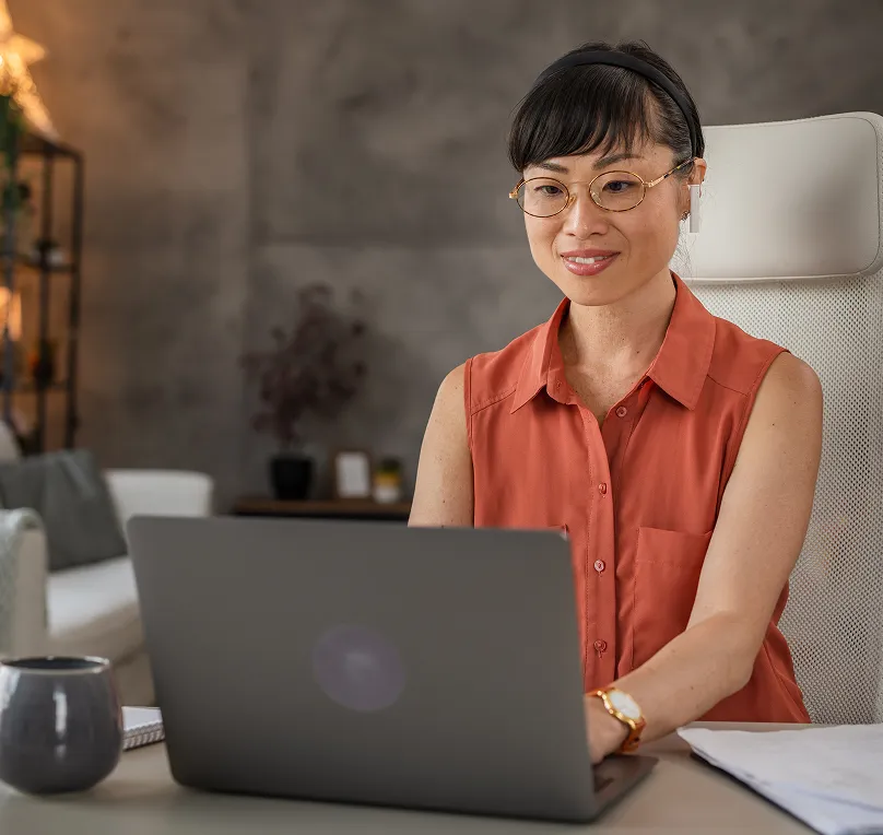 Woman wearing glasses and a sleeveless rust-coloured blouse working on a laptop in a modern home office setting.