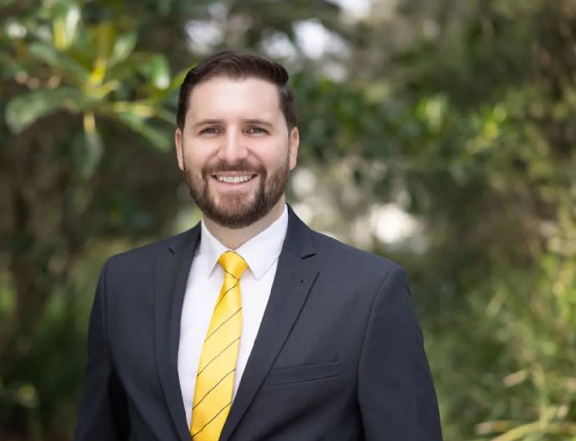 Smiling man in a suit with a yellow striped tie standing outdoors, with greenery in the background.