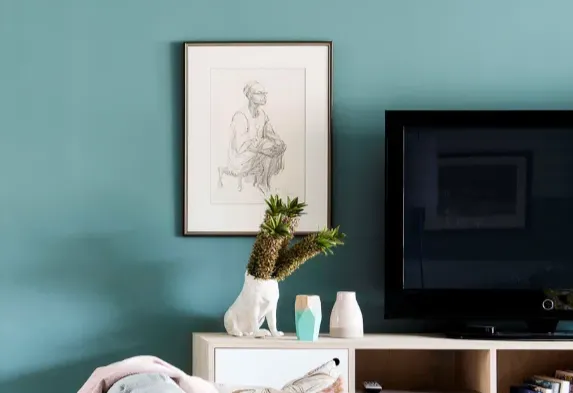 Modern living room interior featuring a teal painted wall, framed sketch artwork, flat-screen TV, and decorative vases on a white entertainment unit.