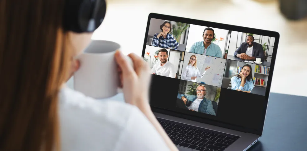 Woman participating in a video conference call while holding a coffee mug, with six diverse colleagues visible on her laptop screen.